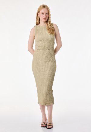 Blondynka stoi z rękami za plecami, ubrana w bezrękawnikową, beżową, teksturowaną sukienkę midi i czarne sandały na ramiączkach na białym tle.