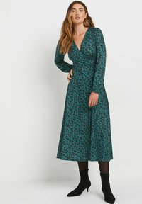 Robe portefeuille teal avec imprimé léopard noir, manches longues, décolleté en V et jupe fluide. Associée à des bottines à talons noirs hauteur cheville.