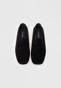Μαύρο σουέτ μοκασίνι με στρογγυλή μύτη, με διακοσμητική λωρίδα και αντίθετη ραφή κατά μήκος των άκρων. Ο λογότυπος της μάρκας ορατός στο εσωτερικό.
