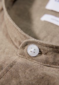 Cuello de camisa de lino beige con un botón blanco de cuatro orificios. Tejido texturizado con detalles de costura visibles. Etiquetas parcialmente visibles.