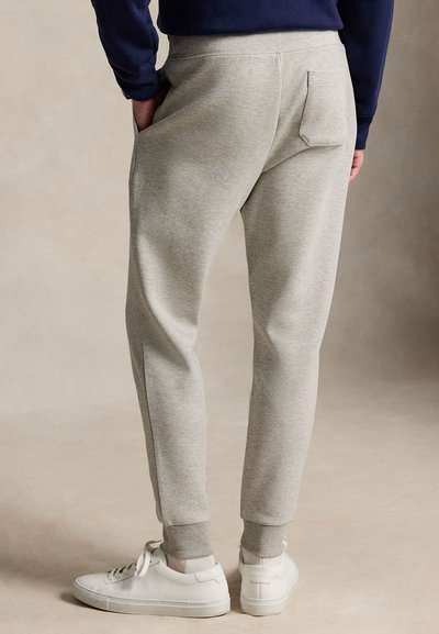 Personne portant un pantalon de jogging gris clair, des baskets blanches et un haut bleu marine, debout avec une main dans la poche sur un fond neutre.