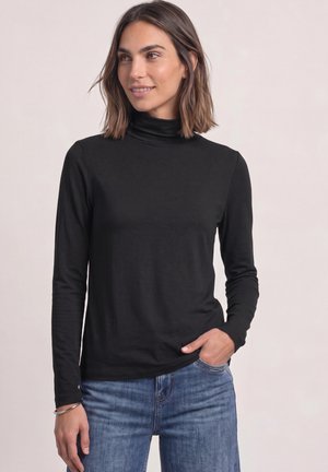 Donna con capelli fino alle spalle che indossa una maglia nera a collo alto con maniche lunghe e jeans blu, in piedi con una mano in tasca.