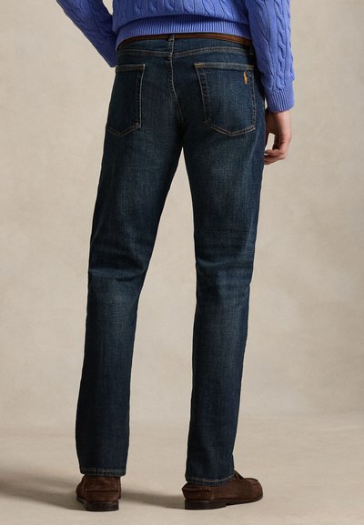 Άνδρας που φοράει σκούρα μπλε τζιν, καφέ ζώνη, μπλε πουλόβερ και καφέ σουέντ παπούτσια, εμφανίζεται από πίσω, στέκεται σε φωτεινό φόντο.