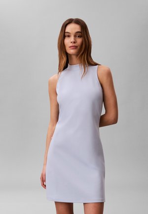 Noorpruunide pikkade juustega noor naine, kes kannab varrukateta kõrge kaelusega helehallit kleiti, seisab lihtsa halli tausta ees.