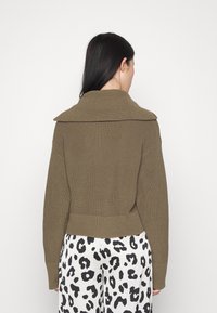 Olivgrüner Strickpullover mit weitem Kragen, strukturiertem Rippmuster und tailliertem Schnitt, kombiniert mit schwarz-weiß gemusterten Leopardenhosen.