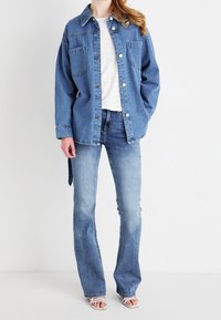 Mujer vestida con una chaqueta de camisa de mezclilla azul sobre una blusa blanca perforada, combinada con jeans acampanados de color azul claro y sandalias blancas con tiras.