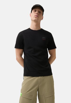 Black cotton t-shirt featuring a small logo patch on the left chest, paired with light khaki cargo shorts with a visible green label.