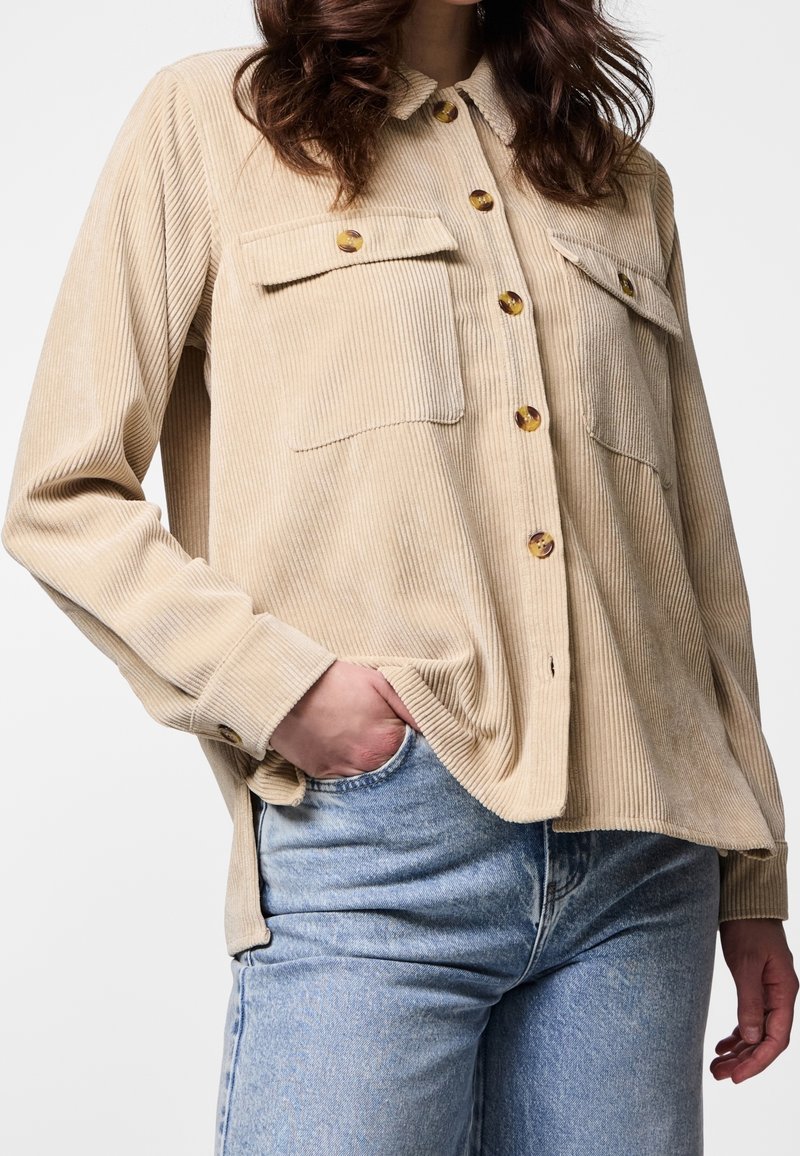 Femme portant une chemise beige en velours côtelé avec poches poitrine et un jean bleu clair, main dans la poche, sur un fond uni.