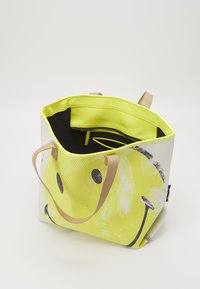 Żółta siatkowa torba tote z wzorem uśmiechniętej buźki. Posiada beżowe uchwyty oraz czarne wnętrze z kieszenią zamykaną na suwak i bocznymi przegródkami.
