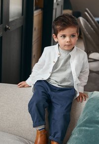 Bambino seduto su un divano grigio chiaro, indossa una camicia bianca abbottonata, una maglietta grigia e pantaloni blu navy con stivali di pelle marrone. Capelli corti e castani.