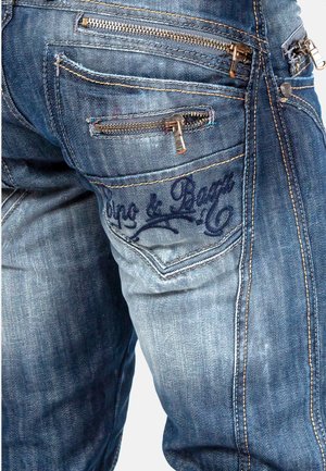 Jeans in denim blu scuro con cuciture evidenti, molteplici tasche con zip e logo ricamato sulla tasca posteriore, che mostrano una finitura texturizzata.