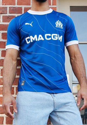 Niebieska piłkarska koszulka z białymi akcentami, z okrągłym dekoltem, teksturowanymi paskami i logo. Noszona z jasnoniebieskimi dżinsami.