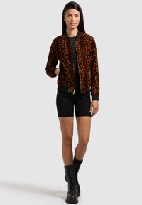 Femeie purtând o jachetă maro cu imprimeu de leopard negru, pantaloni scurți negri și ghete negre cu șireturi, stând în picioare pe un fundal simplu deschis la culoare.