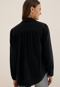 Chemise noire à manches longues avec cuff à boutons, dos plissé et col. Le matériau semble lisse et léger, avec un ourlet incurvé.