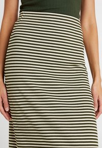 Žebrovaná sukně svislými pruhy v olivově zelené a krémové barvě. Textura je hladká, se střihovým designem přiléhajícím k tělu a s vysokým pasem.