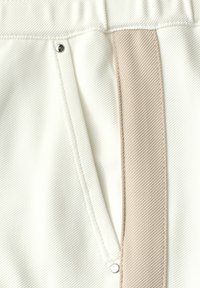 Gros plan d'un tissu blanc texturé avec une rayure verticale beige et deux boutons-pression métalliques argentés sur un vêtement.