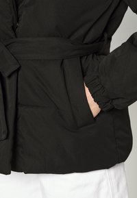 Svart vadderad jacka med bälte i midjan, elastiska ärmslut och sidofickor, tillverkad av ett glatt, matt tyg.