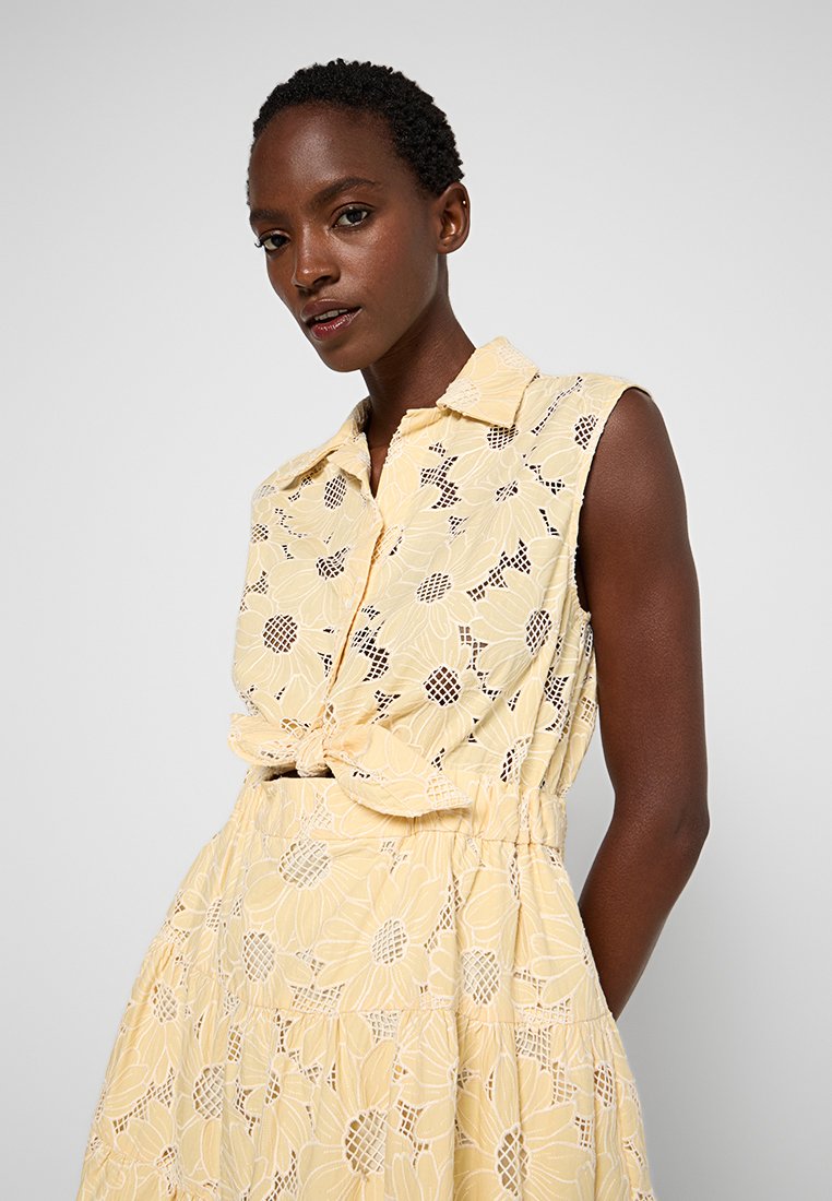 Vestido amarillo con estampado floral, con cuello tipo camisa, sin mangas y cintura ajustable. La tela es ligera y texturizada.