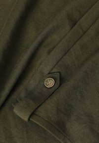Olijfgroene textuurstof met een gladde afwerking, voorzien van een gouden knoop met een decoratief embleem en een gevouwen ontwerp.