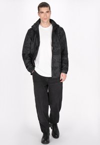 Svart mönstrad jacka med dragkedja fram, hög krage och sidofickor, tillsammans med en vit t-shirt och svarta löst sittande jeans. Svarta stövlar.