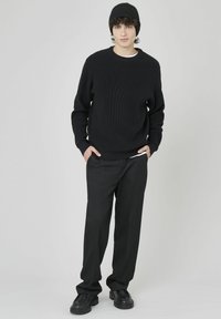 Pull en maille noire avec une texture côtelée, associé à un pantalon noir taille haute et des chaussures noires robustes. Le mannequin se tient avec les mains dans les poches.