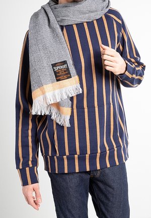 Homme portant un pull à rayures verticales bleu marine et marron avec une poche avant, un jean foncé et une écharpe à chevrons grise avec franges et étiquette.