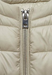 Gros plan sur un tissu matelassé beige avec une grande fermeture éclair métallique et une étiquette "WATER REPELLENT" sur une veste matelassée.