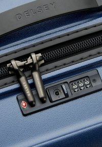 Valigia rigida blu con finitura testurizzata, doppie cerniere nere, lucchetto TSA integrato e etichetta del sistema di sicurezza. Marchio Delsey visibile.