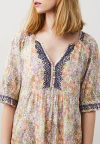 Robe fleurie à manches bouffantes courtes dans des couleurs pastel. Présente un décolleté en V avec un détail de boutons et des accents brodés. Tissu léger.