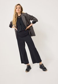Blazer grigio texturizzato su una blusa nera, abbinato a pantaloni neri a gamba larga e sneakers nere; gli accessori includono una collana in oro.