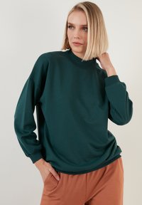 Maglietta verde a maniche lunghe realizzata in materiale morbido, con colletto alto e polsini a costine, dallo stile morbido e abbinata a pantaloni marroni.