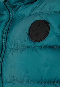 Teal puffer jakna s teksturiranim površinom, s crnim kružnim pečatom s detaljima NMT 97 i prednjim patentnim zatvaračem.