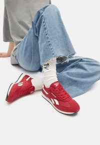 Punased sametist spordijalatsid valgete aktsentide ja paeltega, paigutatud kergelt siniste rebenditega teksade ja valgete sokkide kõrvale, millel on logo.