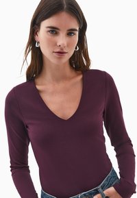 Giovane donna con capelli castani lisci, indossa orecchini a cerchio d'argento, una maglietta a maniche lunghe di un profondo viola con scollatura a V e jeans blu, con una mano in tasca.