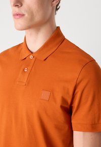 Oransje poloskjorte med klassisk krage, to-knapps knappelukking og strukturert stoff med et firkantet logo-patch på brystet.
