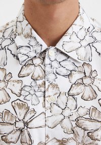 Korte mouwen shirt met een witte achtergrond en grote zwarte en beige bloemenpatronen, knoopsluiting kraag en lichte knoopdetails.