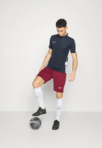 Joven con camiseta negra y blanca de Nike, pantalones cortos burdeos y calcetines blancos equilibrando un balón de fútbol negro de Nike con su pie derecho.