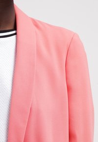 Blazer rosa con tessuto texturizzato, caratterizzato da una chiusura con un solo bottone e rever lisci. Indossato su una maglietta bianca con un motivo a pois.