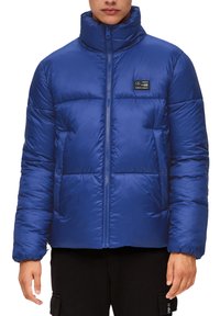 Blauwe pufferjack met hoge kraag, ritssluiting, gewatteerd ontwerp en logopatch op de linkerborst. Gemaakt van een glanzende, gladde stof.