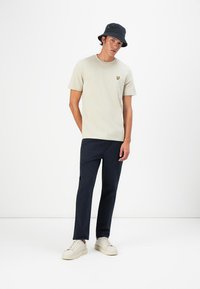 Lys grå t-shirt med lille gul logo, kombineret med marineblå bukser og cremefarvede sneakers. Modellen bærer en sort bucket hat.