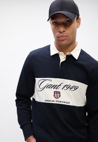 Polo din bumbac navy cu dungi orizontale albe, brodat "Gant 1949" și logo cu scut. Prezintă un guler clasic și mâneci lungi.