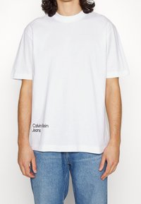 Biała bawełniana t-shirt z okrągłym dekoltem i krótkimi rękawami, z czarnym logo "Calvin Klein Jeans" na dolnej lewej stronie.