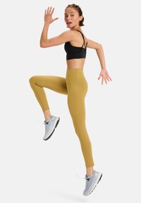 Soutien-gorge de sport noir, leggings moutarde et baskets grises claires. Les leggings sont à taille haute, tandis que les chaussures ont une semelle texturée.