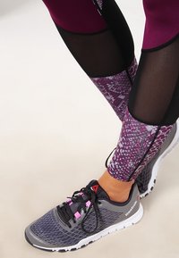 Legginsy w burgundzie i czarnym siatkowym materiale z fioletowym i szarym wzorem. Noszone z szarymi butami sportowymi z różowymi akcentami i czarnymi sznurowadłami.