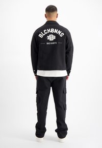 Zwarte denimjack met witte geborduurde tekst op de rug. Gestyled met losse zwarte cargo-broeken en witte schoenen van textuurstof.