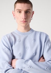 Sweatshirt bleu clair avec un motif de logo texturé, col rond, poignets côtelés et coupe décontractée. Le tissu semble doux et confortable.