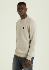 Maglione in beige chiaro con maniche lunghe, colletto rotondo e polsini a coste. Presenta una piccola etichetta logo nera sul petto.
