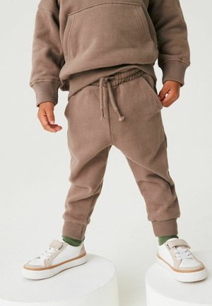 Enfant portant un pantalon de survêtement marron et un sweat à capuche, avec des chaussettes vertes et des baskets blanches, debout sur deux plateformes cylindriques blanches.