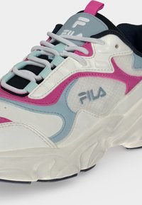 Λευκό, ροζ και μπλε αθλητικό παπούτσι FILA με χοντρή σόλα, γκρι κορδόνια και λογότυπο στο πλάι και στη γλώσσα, εμφανιζόμενο σε γωνιακή κοντινή λήψη.