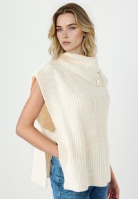 Poncho lavorato a maglia color crema con texture a coste, collo ampio a scialle e dettagli con bottoni sulle spalle; presenta spacchi laterali e una vestibilità comoda.